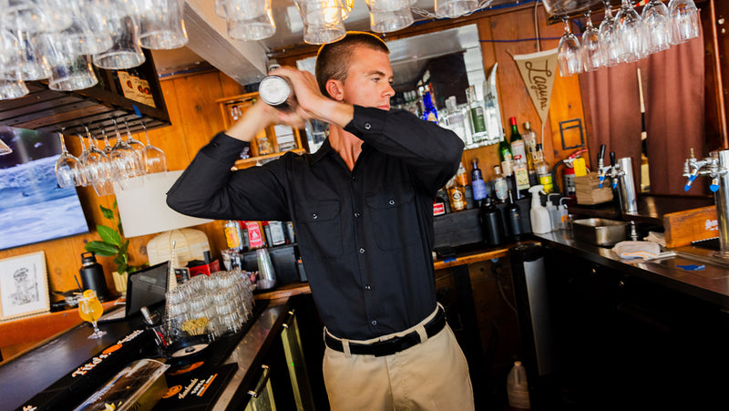 A bartender in a black long-sleeve button-up shirt and khaki pants shakes a cocktail shaker behind a bar counter with hanging glassware and bottles in the background.