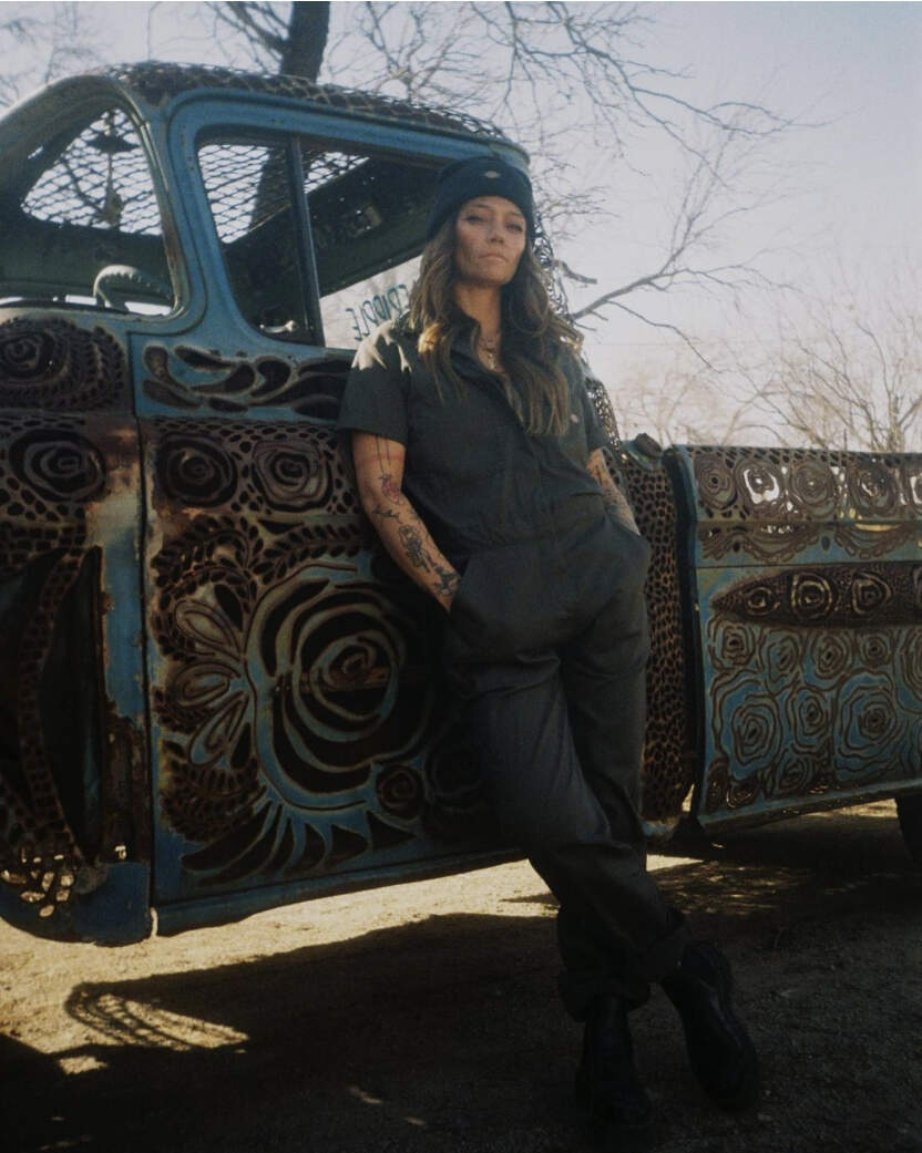 Rae Ripple leaning on vintage decorated truck wearing warming beanie and short sleeve coveralls