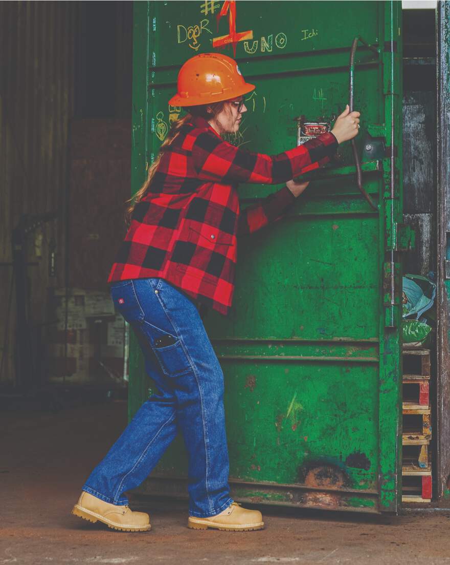 Woman in hardhat wearing plaid top and carpenter jeans opening metal door