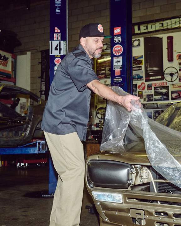 Mechanic wearing blue Dickies button down and Khakis pulling a plastic tarp over an antique car.