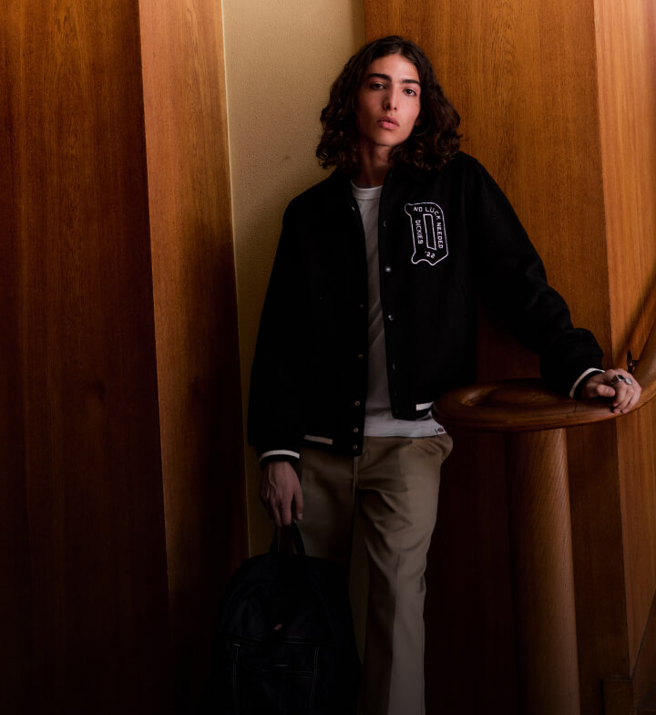 A person wearing a black collegiate jacket standing next to a wall looking at the camera.