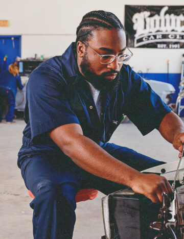 A man wearing dickies coveralls working under the hood of a vehicle.