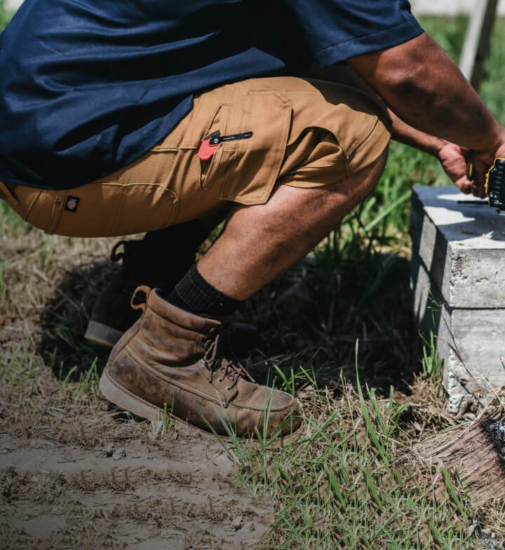 A worker squatting while using an electric tool and wearing khaki-colored shorts.