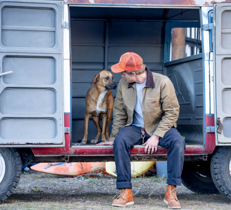 A man sitting on the outside of a van next to a dog.