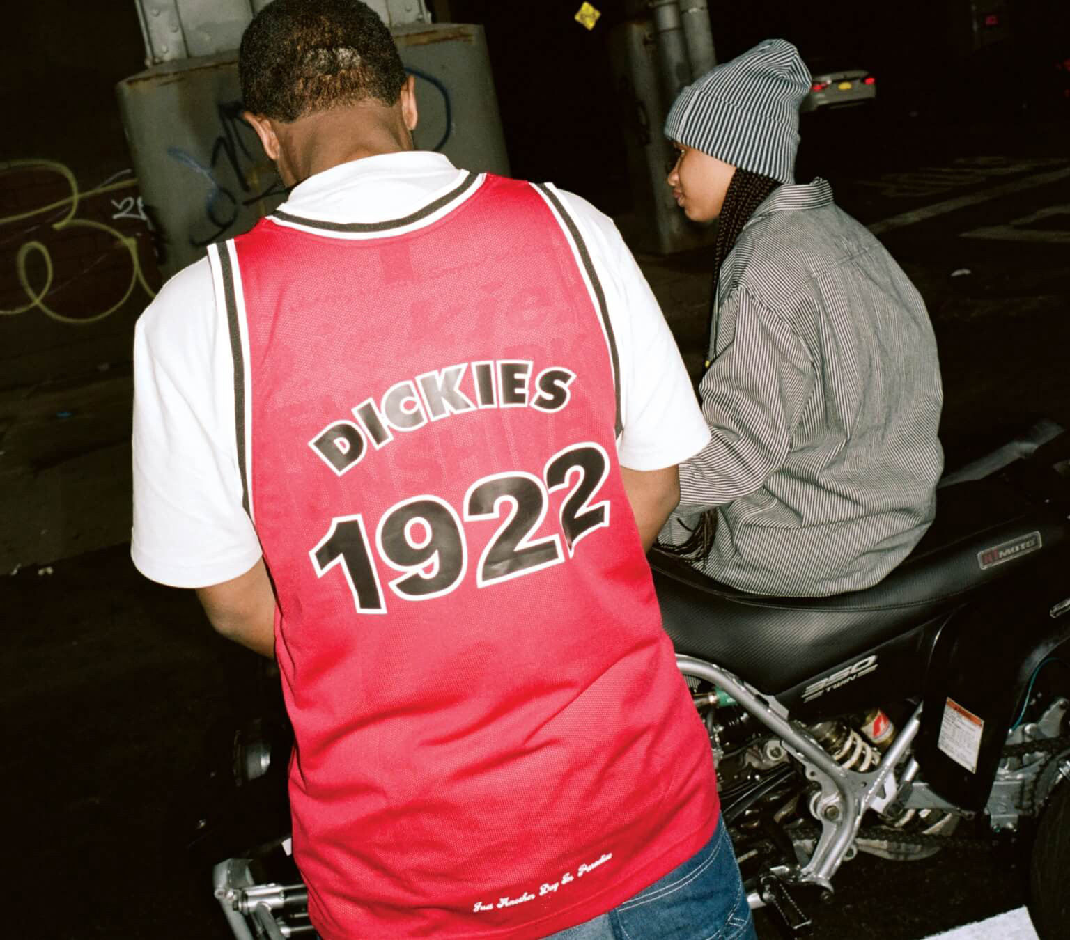 A man wearing a dickies 1922 jersey and a women sitting on a quad vehicle
