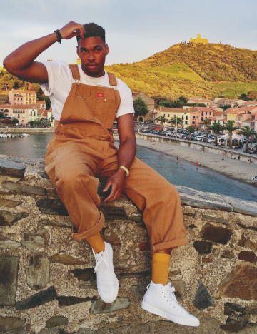 A man sitting on a rock ledge wearing duck colored bib overalls.