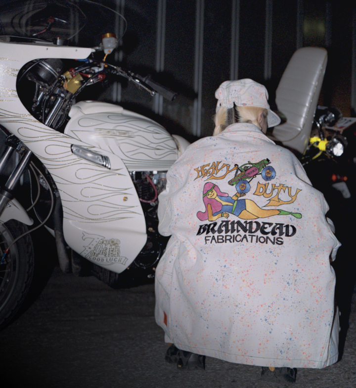 A person wearing a white brain dead jacket crouching next to a white motorcycle.