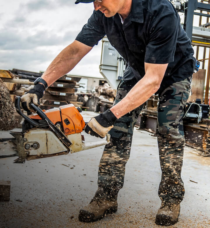 A worker using a chainsaw with sawdust flying on their camo cargo pants.
