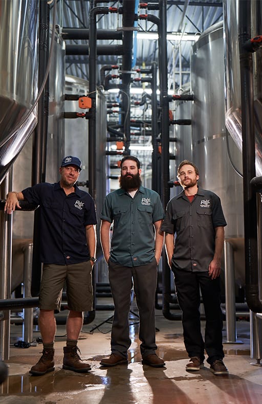 Three brewers posing next to fermenting tanks.
