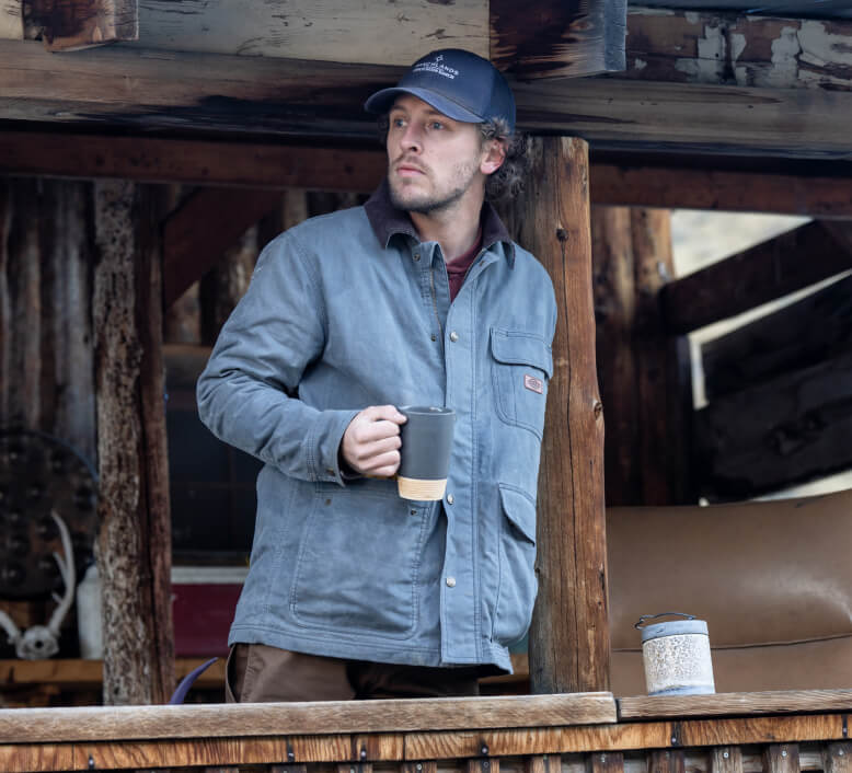 A man standing outside wearing a blue chore waxed jacket leaning on a wooden pole.